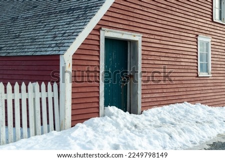A closed small vintage green wooden shed door with a lock and latch. The barn has a vibrant red clapboard exterior wall with horizontal lines, white trim, and peeling paint with a mound of fresh snow.