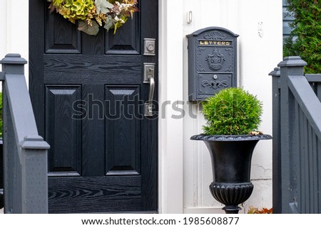 A closed solid wood black front door of a house. There's a black mailbox hanging on the white exterior wall. There's a flower pot with a green hedge. The door has a colorful flower wreath. 