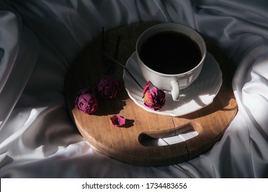 A Closep Of Porcelain Cup Of Coffee On Wooden Cutting Board And Some Dry Roses On Silk Sheet . Coffee To Bed .  Parisian Style . 