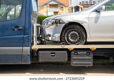 A closer view of the front-left side of a silver car with collision damage and a dented bumper strapped on a tow truck platform in front of a residential neighborhood