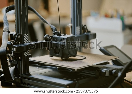 Close-up of a 3D printer during operation in a workshop setting creating an object with detailed components visible and equipment in the background.