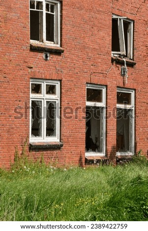 Closeup of an abandoned and dilapidated farm house with red bricks and broken glass on a neglected terrain with tall grass. Concept of rural depopulation.
