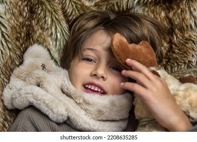 Close-up Of An Adorable Smiling Child Looking At The Camera On A Brown Blanket. Child Inside The House On A Cold Winter Day.  