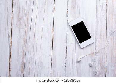 Closeup Aerial View Of White Smart Phone With Black Isolated Screen And Headphones On White Wooden Surface. Mockup. Copy Space Available.