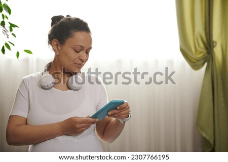 Close-up African American adult pregnant woman with headphones, using smartphone, adjusting soundtrack in mobile app with pregnancy yoga , ready to practicing stretching exercises at home interior