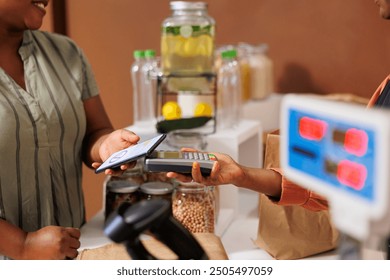 Closeup of african american client using mobile device on pos machine to pay for fresh organic product at local grocery store. Black woman making nfc payment with cell phone for bio food items. - Powered by Shutterstock