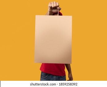 Closeup Of African American Delivery Man In Red Uniform Holding And Showing Craft Paper Bag To Camera, Isolated On Orange Background. Design, Mockup, Blank Space On Package For Advert