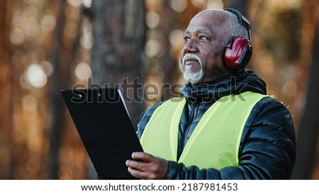 Close-up african american male professional foreman forestry engineer standing outdoors wearing protective headphones assessing situation making tree felling plan writing data reforestation projectt