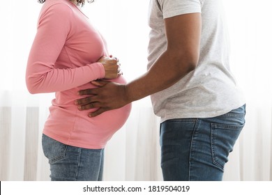 Closeup of african american pregnant couple embracing at home. Unrecognizable black husband and expecting wife with big belly standing by window and hugging, pregnancy, family and marriage concept