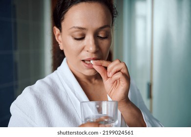 Close-up Of African American Pretty Woman, Wearing White Bathrobe, Holding A Glass Of Water And Taking Nutritional Supplement In The Morning. Health Care Concept. Beauty Treatment. Weight Loss Capsule