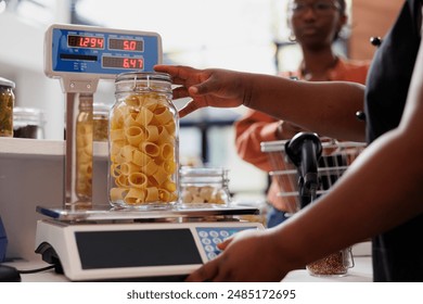 Closeup of african american vendor using digital scale to measure weight of fresh organic product in glass container. Detailed view of merchant weighing jar of bio pasta while client waits at counter. - Powered by Shutterstock