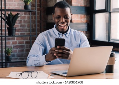 Closeup of African guy sitting at table in office and smiling happily while holding smartphone