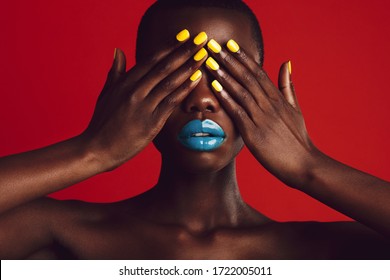 Close-up of african woman with colorful makeup covering her eyes with her hands. Female model with vibrant makeup on red background.