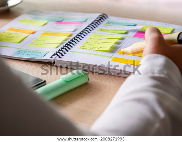 Close-up of agenda organize with color-coding\
sticky notes for time management. Productive schedule for\
appointments and reminders. Hand holding a yellow highlighter\
marker. Organization and\
planning