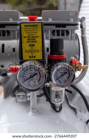 Close-up of an air compressor pressure switch assembly, featuring a black control box with a red emergency stop button, dual analog pressure gauges, and a red output pressure regulator knob