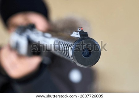 Close-up of air rifle muzzle and front fiber optic sight aiming at target. Focused barrel detail against blurred young man shooter background with precision sports equipment hunting and shooting.
