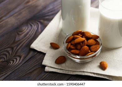Close-up of almond milk in a glass mug and a refillable glass bottle, next to it lie almonds and a textile napkin on a wooden table.