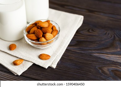 Close-up of almond milk in a glass mug and a refillable glass bottle, next to it lie almonds and a textile napkin on a wooden table.