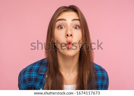 Closeup of amazed childish cute girl in checkered shirt standing with pout lips making fish face, having fun, showing surprised silly expression. indoor studio shot isolated on pink background
