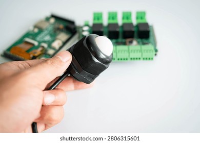 Close-up of an ambient light sensor held by hand with an electronic controller board in the background, representing IoT, smart building automation, environmental monitoring, embedded systems, energy management and industrial technology.
