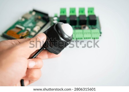 Close-up of an ambient light sensor held by hand with an electronic controller board in the background, representing IoT, smart building automation, environmental monitoring, embedded systems, energy management and industrial technology.