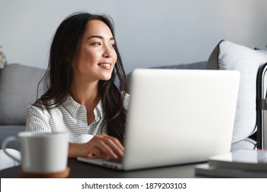 Close-up Of Ambitious Beautiful Asian Female Employee Working From Home. Girl On Remote Using Laptop, Smiling Dreamy While Looking Outside Window. Student Feel Inspired Writing Essay.