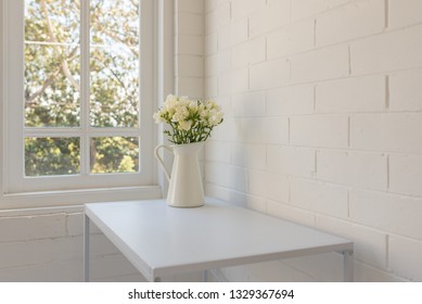 Closeup angled view of white freesias in jug on table against window and painted brick wall (selective focus)
