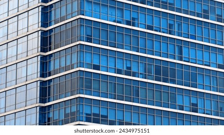 A close-up, angular view of a modern skyscraper featuring a vast grid of blue glass windows. The architectural design emphasizes clean horizontal and vertical lines, creating a repetitive and symmetrical geometric pattern across the building's facade. Light reflections on the glass panels and the varying shades of blue provide a sense of depth and transparency to the massive urban structure.