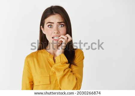 Close-up of anxious young woman biting fingernails, staring at camera insecure and nervous, standing unconfident on white background