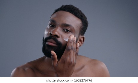 Close-up of appealing nude black male model isolated on grey background. Portrait of confident african man applying cream on his face looking in the mirror.