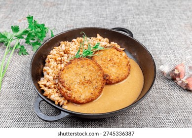 Close-up of appetizing fried fish cutlets made from carp with loose spelt porridge and rich gravy (sauce), served in a traditional cast iron skillet on a textured tablecloth. The dish is garnished wit