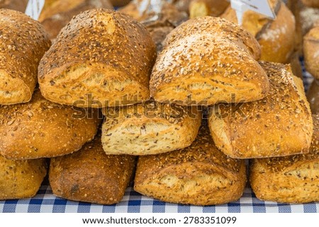 A close-up and appetizing photograph of a generous stack of rustic multigrain bread rolls piled on a blue and white checkered tablecloth at an outdoor French market. The rolls are baked to a deep golden-brown and generously coated with a mixture of sesame and flax seeds that stud the entire crust surface, creating a richly textured and visually appealing exterior. Where one roll is turned to reveal its interior, a dense, moist crumb with embedded seeds is visible, suggesting a hearty, nutritious loaf. The informal stacking of the rolls adds to the authentic, abundant character of the market display. Additional bread varieties are faintly visible in the soft-focus background. The warm, inviting tones and artisan quality of the baked goods evoke the atmosphere of a traditional French farmers market or village fair. This image suits artisan bakery and delicatessen marketing, organic and wholefood brands, farmers market and local produce content, food tourism, healthy eating and nutrition, gastronomy and culinary editorial, and southern France regional lifestyle campaigns.