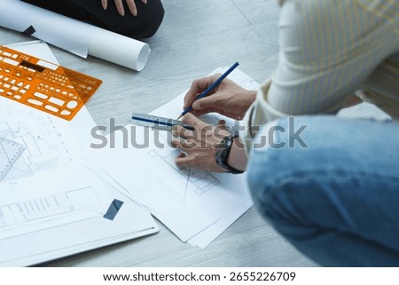 Close-up of architect drawing structural plan using pencil and ruler on blueprint sheet during design process on wooden floor.