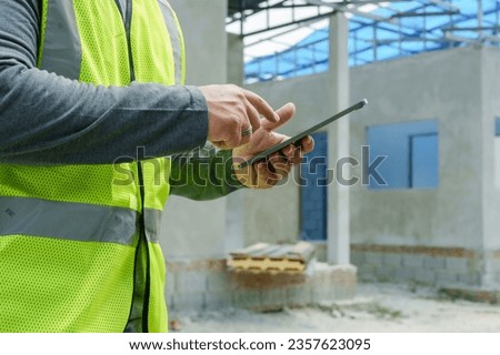 Close-up. Architect's hand checking construction plan from tablet. Get home decor planned quickly. Asian man wearing green reflective vest and safety helmet Stand outside the house under construction