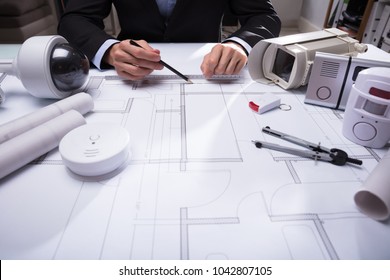 Close-up Of An Architect's Hand Drawing Blueprint With Security Equipments On Desk