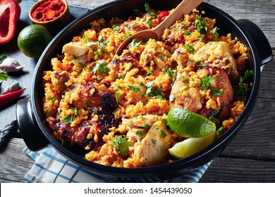 close-up of Arroz con pollo, spanish cuisine, rice with chicken and veggies in a black saucepan on a wooden rustic  table with ingredients on a slate board, view from above