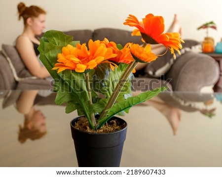 Close-up of artificial orange flowers in pot on glass coffee table in front of blurred figure of young woman lying on sofa