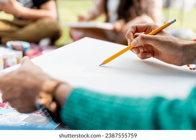 A close-up of an artist's hand holding a pencil and starting to sketch on a blank canvas, with art supplies and a park setting in the background - Powered by Shutterstock