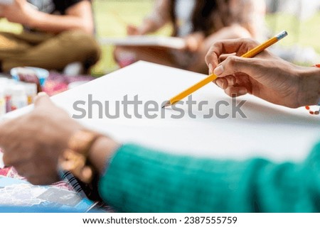 A close-up of an artist's hand holding a pencil and starting to sketch on a blank canvas, with art supplies and a park setting in the background