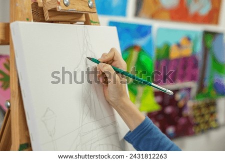 Close-up of an artist's hand holding pencil, drawing preliminary lines for sketch on white canvas with colorful paintings in background.