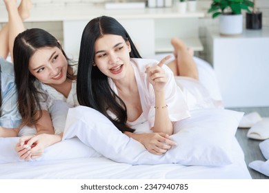 Closeup Asian beautiful cheerful happy female girlfriends in casual pajamas outfit laying lying down on pillows smiling talking having conversation slumber party together at night on bed in bedroom.