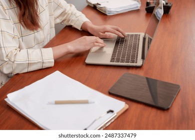 Close-up of an Asian female office worker typing on a laptop at a wooden desk. Documents, pen, and tablet are also placed on the desk. Modern office setting with focus on her hands.