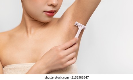 Closeup Of Asian Female Shaving Armpits Removing Underarms Hair With Safety Razor In Bathroom, Standing Wrapped In Towel, White Studio Background. Armpit Depilation Routine, Body And Skin Care Concept