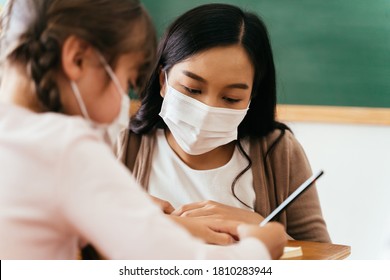 Close-up Of Asian Female Teacher Wearing A Face Mask In School Building Tutoring A Primary Student Girl. Elementary Pupil Is Writing And Learning In Classroom. Covid-19 School Reopen Concept