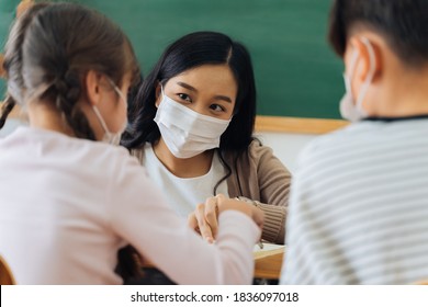 Close-up of Asian female teacher wearing a face mask in school building tutoring a primary student children. Elementary pupils writing and learning in classroom. Covid-19 school reopen concept