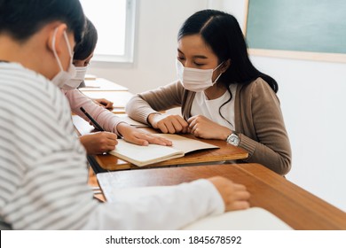 Close-up Of Asian Female Teacher Wearing A Face Mask In School Building Tutoring A Primary Student Children. Elementary Pupils Writing And Learning In Classroom. Covid-19 School Reopen Concept