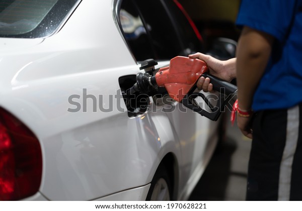 Close-up of Asian gas station worker\'s hand in\
blue uniform, holding a fuel nozzle filling fuel into a white car\
tank that park for refueling in a gas station at night.\
Transportation Energy\
concept.