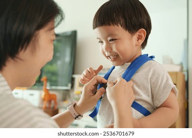 Close-up of Asian mother fastening a blue backpack on a smiling young boy, creating a warm and joyful moment of bonding in a bright living room - Powered by Shutterstock