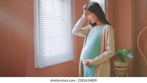 closeup asian pregnant woman standing by the window having headache and worried - Powered by Shutterstock