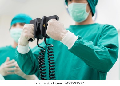 Close-up of Asian surgeon in green scrubs holding defibrillator paddles in operating room. Emphasis on emergency care, precision, and advanced medical equipment in sterile environment in hospital - Powered by Shutterstock